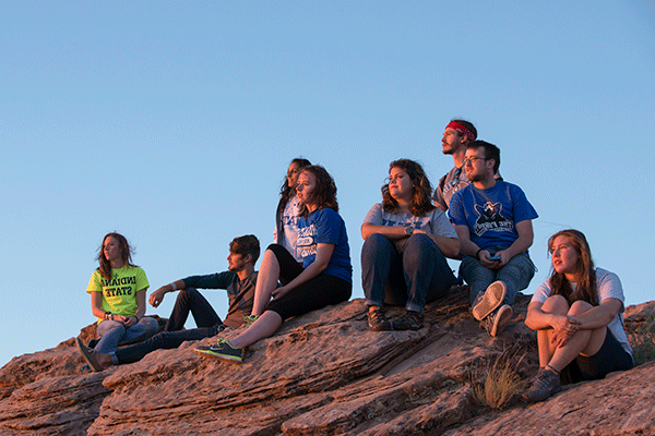 Eight Honors students, of mixed races, sit on a canyon rock, looking to the right. They wear a variety of blue, white, grey, and green clothing.